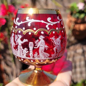 May include: A ruby red glass with a gold rim and base. The glass is decorated with white etched figures and floral designs. The glass is likely a decorative piece or a unique drinking vessel.