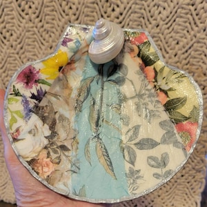 May include: A decorative seashell-shaped trinket dish. The dish is adorned with a floral pattern in shades of pink, yellow, and blue, with a silver trim. A small, pearlescent seashell is attached to the top.