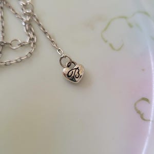 May include: A silver-toned necklace with a heart-shaped pendant. The pendant features the letter "B" in a decorative script. The necklace is resting on a light-colored surface.