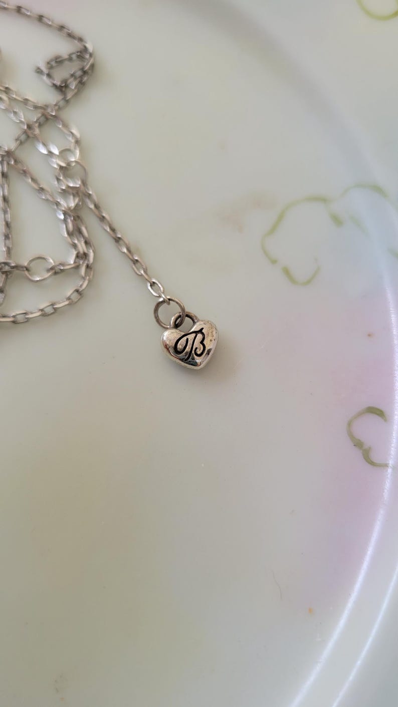 May include: A silver-toned necklace with a heart-shaped pendant. The pendant features the letter "B" in a decorative script. The necklace is resting on a light-colored surface.