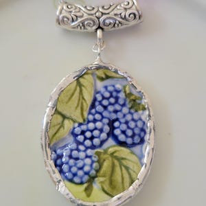 May include: An oval pendant necklace featuring a ceramic shard with a blue blackberry and green leaf pattern. The pendant is encased in a silver-toned metal frame with a textured bail and hangs from a silver-toned chain link necklace.