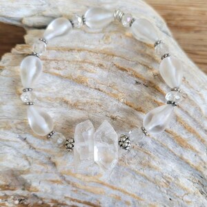 May include: A stretch bracelet featuring alternating frosted white teardrop beads and clear faceted crystal beads. The design includes two central clear quartz points flanked by small silver-toned metal spacer beads, creating a circular arrangement of translucent and opaque elements.