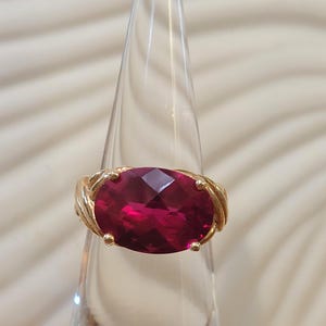 May include: A gold ring featuring a large, faceted, oval-shaped, deep red gemstone. The ring's band has a textured, twisted design. The gemstone is held in place by four small gold prongs.