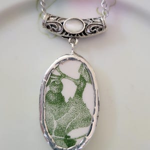 May include: An oval pendant necklace featuring a white ceramic shard with a green botanical print, encased in a silver-toned metal frame. The pendant hangs from a silver-toned chain and includes a decorative bail with an inlaid white cabochon.