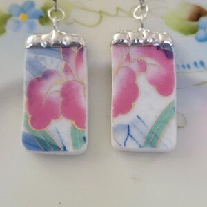 May include: A pair of rectangular dangle earrings featuring white ceramic shards with a pink floral pattern, green leaves, and gold-toned outlines. The ceramic pieces are capped with silver-toned metal hardware and attached to silver-toned earring hooks.