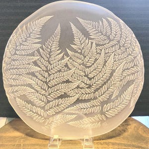 May include: A circular, frosted glass plate featuring an intricate, etched design of multiple fern fronds across its surface. The translucent glass displays detailed leaf patterns with a matte finish, and a small square maker's mark is visible.