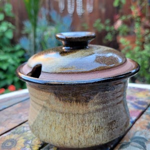 May include: A handcrafted ceramic sugar bowl with a lid. The bowl has a brown and tan glaze with a textured appearance. The lid features a small knob and a slot for a spoon. The sugar bowl is a functional and decorative kitchen item.