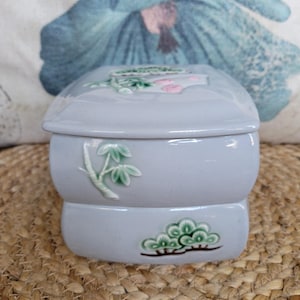 May include: A small, gray ceramic box with a lid. The box is decorated with green and pink floral designs. The box is sitting on a woven surface.