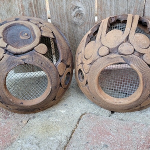 May include: Two spherical brown clay sculptures featuring intricate cut-out patterns and circular openings. Each piece is constructed with textured, layered clay elements and integrated metal wire mesh backing, creating a hollow, dome-like structure.