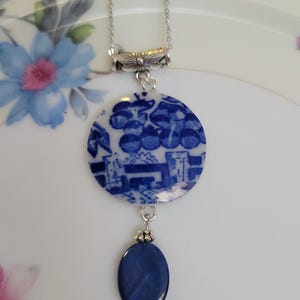 May include: A silver chain necklace featuring a circular pendant made from blue and white patterned ceramic shard, accented with a smaller oval-shaped dark blue bead and silver-toned metal findings at the top and bottom of the bead.