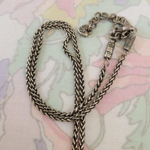 Puede incluir: Collar plateado con un dise&ntilde;o de cadena trenzada. El collar presenta un cierre en forma de coraz&oacute;n y cuentas cil&iacute;ndricas decorativas. La cadena se muestra sobre un fondo con estampado floral.