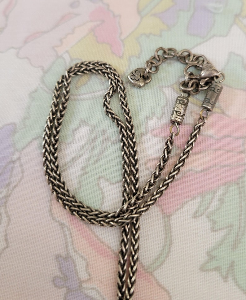 Puede incluir: Collar plateado con un dise&ntilde;o de cadena trenzada. El collar presenta un cierre en forma de coraz&oacute;n y cuentas cil&iacute;ndricas decorativas. La cadena se muestra sobre un fondo con estampado floral.