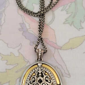 Puede incluir: Collar plateado con un colgante ovalado. El colgante tiene un borde dorado, un dise&ntilde;o de filigrana plateada y est&aacute; incrustado con peque&ntilde;os cristales transparentes. La cadena del collar es de dise&ntilde;o tejido.