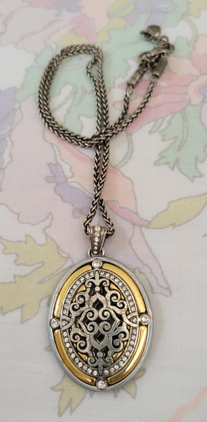 Puede incluir: Collar plateado con un colgante ovalado. El colgante tiene un borde dorado, un dise&ntilde;o de filigrana plateada y est&aacute; incrustado con peque&ntilde;os cristales transparentes. La cadena del collar es de dise&ntilde;o tejido.