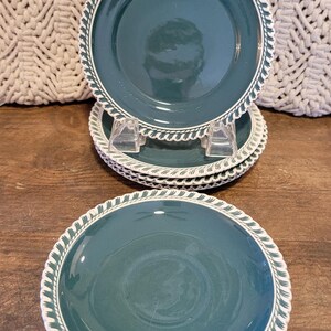 May include: A stack of teal-colored plates with a white and green decorative border. The plates are arranged on a wooden surface, with a textured off-white background visible in the upper portion of the image. The plates have a classic design.