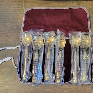 May include: A set of six metallic spoons with dark-toned handles, each individually wrapped in clear plastic sleeves. The spoons are arranged side-by-side within a deep burgundy velvet roll-up storage case featuring white fabric ties.