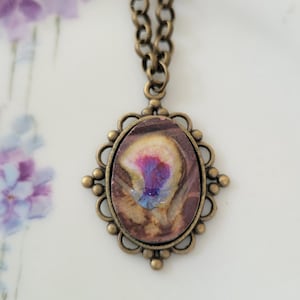 May include: An oval pendant necklace featuring a bronze-toned metal setting with a decorative filigree border. The center inlay displays a marbled pattern in shades of brown, cream, and a vibrant purple-blue focal point, suspended on a bronze chain.