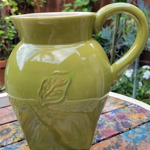 May include: A green ceramic pitcher with a leaf design. The pitcher has a rounded body, a curved handle, and a square base. The leaf design is embossed on the front. The pitcher is sitting on a wooden table.