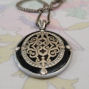 Puede incluir: Collar plateado con un colgante redondo. El colgante presenta un centro de esmalte negro con un dise&ntilde;o de filigrana plateada, rodeado de cristales transparentes. La cadena es de estilo tejido.