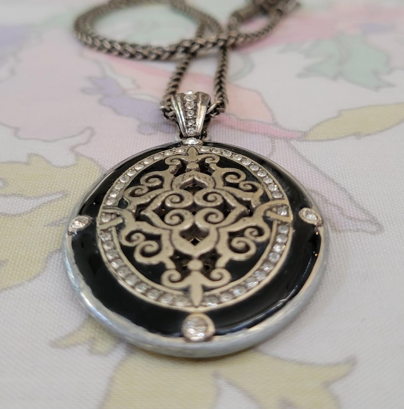 Puede incluir: Collar plateado con un colgante redondo. El colgante presenta un centro de esmalte negro con un dise&ntilde;o de filigrana plateada, rodeado de cristales transparentes. La cadena es de estilo tejido.