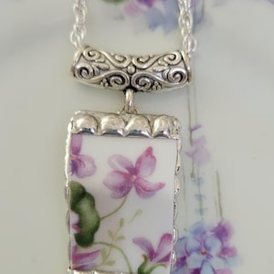May include: A rectangular pendant necklace featuring a piece of white ceramic with a purple and green floral pattern. The ceramic is set in a scalloped silver-tone metal frame with a decorative, embossed silver-tone bail attached to a silver chain.