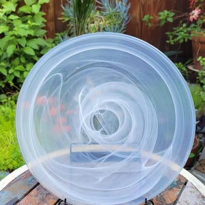 May include: A circular glass plate featuring a translucent finish with swirling white marbled patterns throughout the surface. The plate is held upright by a black metal display stand, showcasing the intricate, cloud-like white streaks.