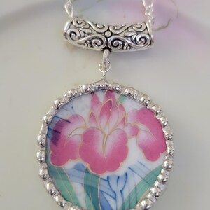 May include: A circular pendant necklace featuring a pink iris flower illustration on a white background, encased in a silver-toned beaded metal frame. The pendant hangs from a silver-toned chain by a decorative, scroll-patterned metal bail.