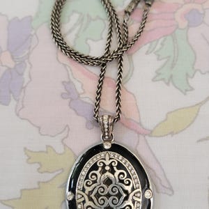 Puede incluir: Collar plateado con un colgante ovalado. El colgante tiene un borde de esmalte negro, un dise&ntilde;o de filigrana plateada y est&aacute; adornado con cristales transparentes. La cadena es de estilo trenzado.