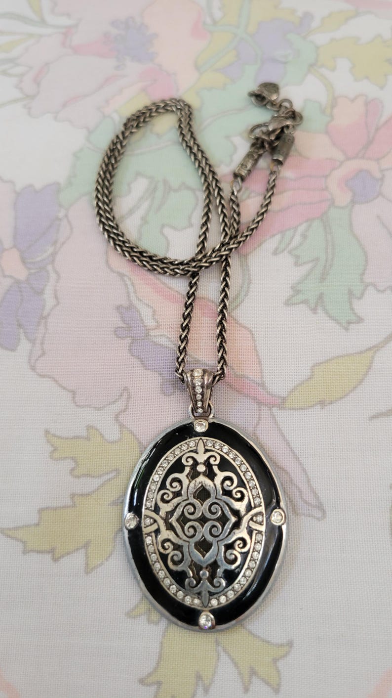 Puede incluir: Collar plateado con un colgante ovalado. El colgante tiene un borde de esmalte negro, un dise&ntilde;o de filigrana plateada y est&aacute; adornado con cristales transparentes. La cadena es de estilo trenzado.