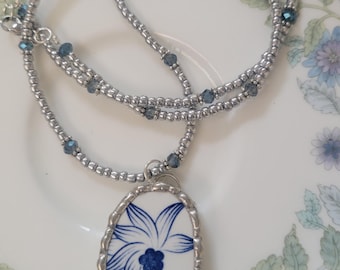 May include: A handmade necklace featuring a pendant made from a piece of porcelain with a blue floral design. The pendant is framed in silver and hangs from a silver beaded chain with blue accents. The necklace rests on a white plate with blue floral patterns.