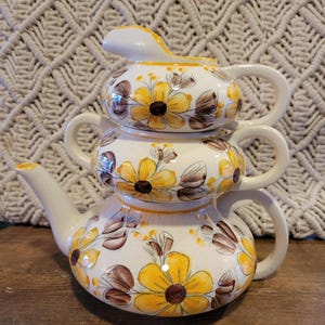 May include: A set of three ceramic teapots with a floral design. The teapots are stacked on top of each other, with the smallest teapot on top. The teapots are decorated with yellow and brown flowers on a white background.