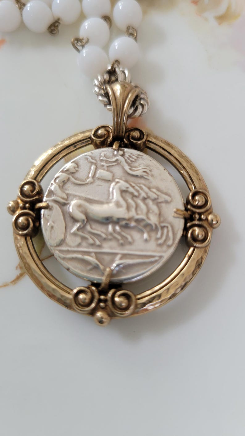 May include: A vintage necklace featuring a silver pendant with a chariot scene and a gold-toned frame. The pendant is attached to a strand of white, round beads. The necklace has a classic, antique aesthetic.