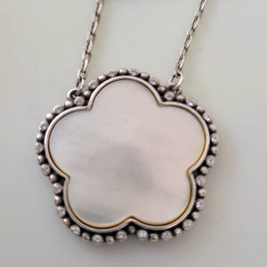 May include: Silver-tone necklace featuring a clover-shaped pendant. The pendant has a mother-of-pearl center, surrounded by a beaded border and small clear stones. The chain is delicate and has a simple design.