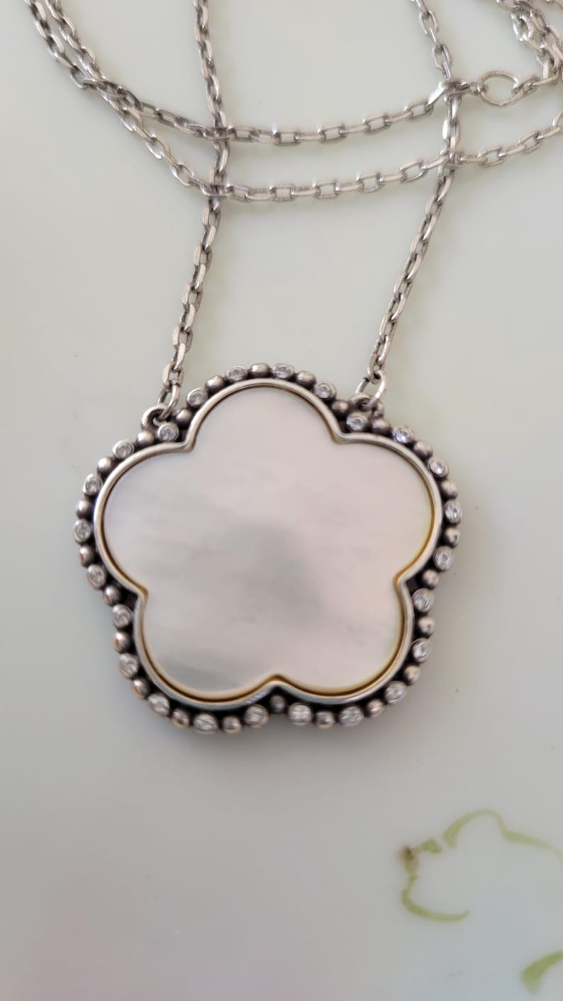 May include: Silver-tone necklace featuring a clover-shaped pendant. The pendant has a mother-of-pearl center, surrounded by a beaded border and small clear stones. The chain is delicate and has a simple design.