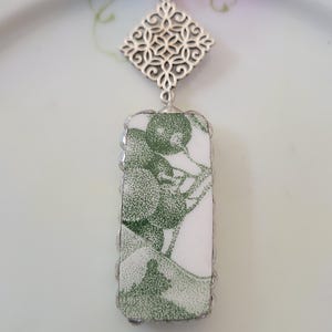 May include: A rectangular pendant necklace featuring a white ceramic shard with a green stippled botanical print. The shard is set in a silver-toned metal frame with a decorative filigree diamond-shaped bail attached to a thin silver-toned chain.