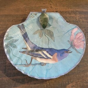 May include: A decorative, shell-shaped pendant with a painted bird design. The pendant features a blue bird with a pink chest and a green gemstone at the top. The background is a light blue with floral accents and a silver border.