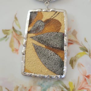May include: A rectangular pendant necklace featuring a gold and gray leaf-patterned inlay encased in a silver-toned metal frame with a decorative filigree bail. The pendant hangs from a silver-toned chain, showcasing a textured, metallic finish.