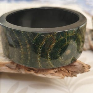 May include: A wide, cylindrical bangle bracelet featuring a polished black interior and a dark green exterior surface. The outer band is decorated with a repeating pattern of golden-brown, circular, sunburst-like radial line designs.