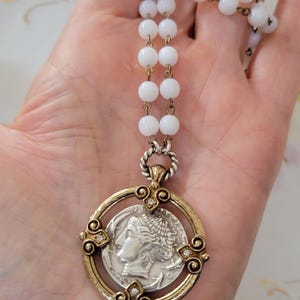 May include: A vintage necklace featuring a silver coin pendant with a profile image, set in a gold-toned frame with clear accents. The necklace has a strand of white, round beads and gold-toned chain links.