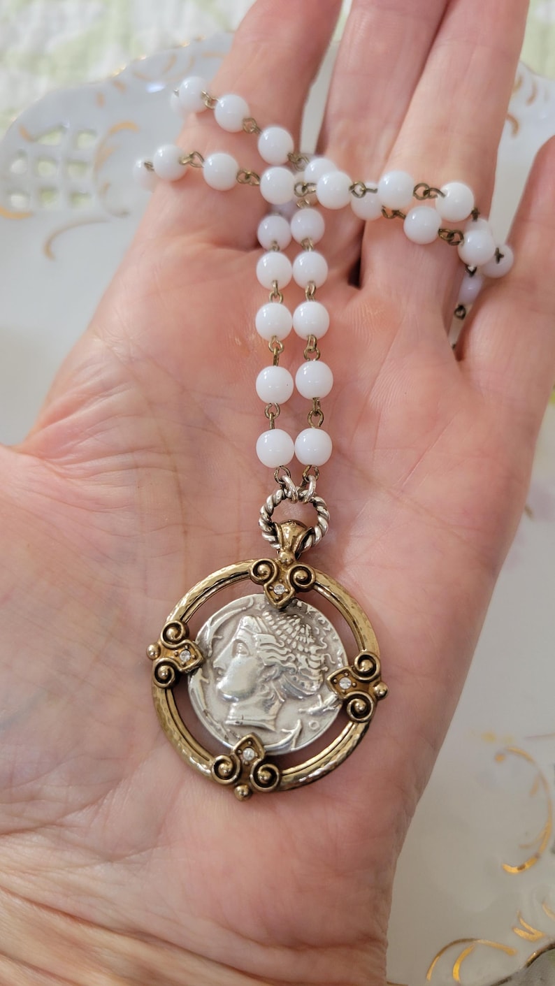 May include: A vintage necklace featuring a silver coin pendant with a profile image, set in a gold-toned frame with clear accents. The necklace has a strand of white, round beads and gold-toned chain links.