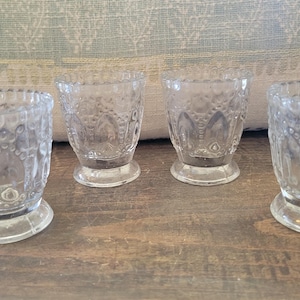 May include: A set of four clear glass egg cups featuring a textured hobnail pattern and a scalloped rim. Each cup has a sturdy circular base and a tapered body with embossed decorative details around the exterior surface of the glass.