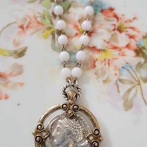May include: A white beaded necklace with a pendant featuring a silver coin with a profile image. The pendant is set in a gold-toned frame with decorative accents and small clear stones. The necklace is displayed on a floral patterned surface.