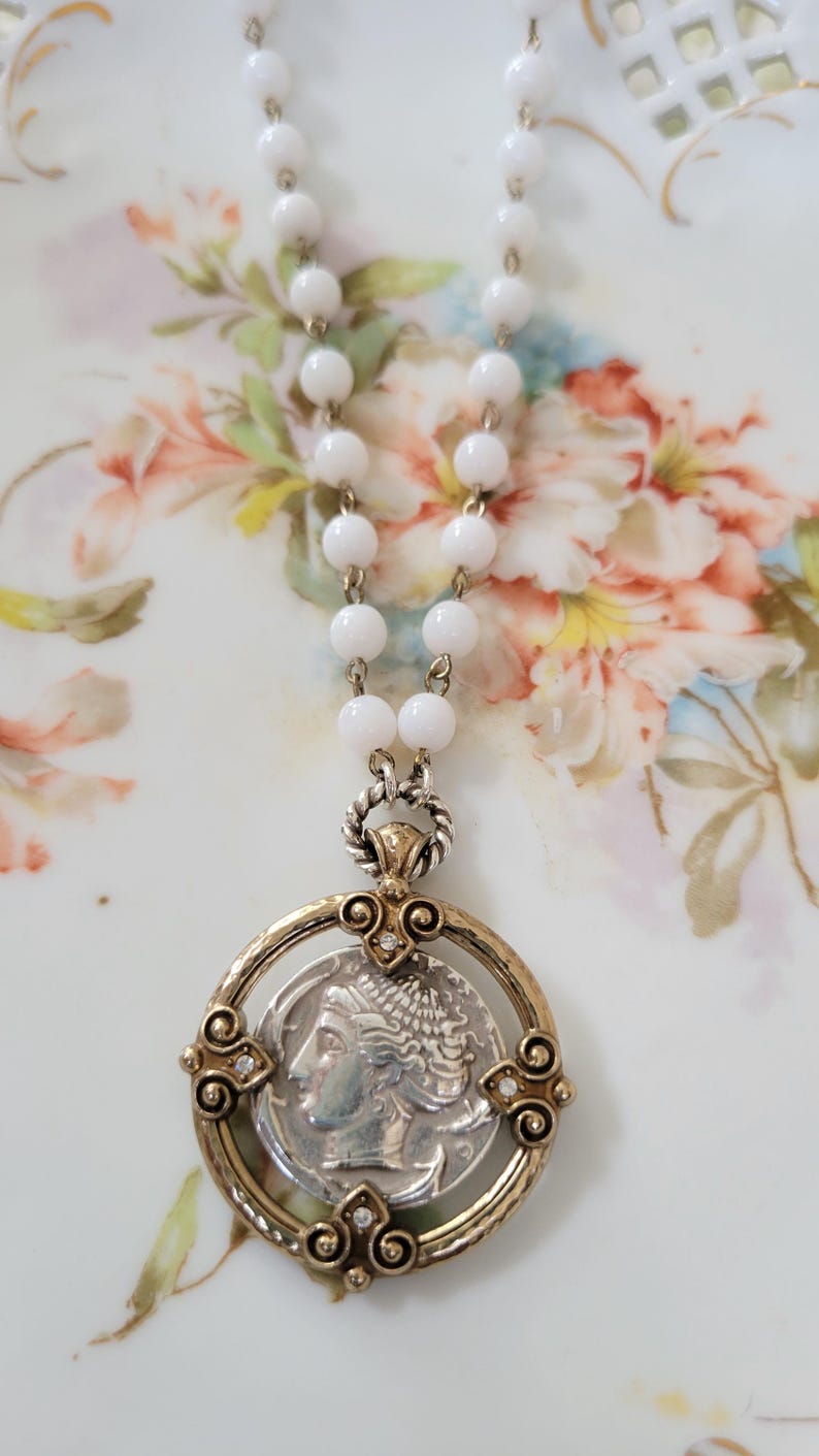May include: A white beaded necklace with a pendant featuring a silver coin with a profile image. The pendant is set in a gold-toned frame with decorative accents and small clear stones. The necklace is displayed on a floral patterned surface.