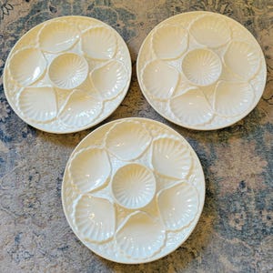May include: Three white ceramic oyster plates. Each plate features a central well and six shell-shaped compartments. The plates have a scalloped edge design. The plates are arranged on a patterned surface.