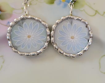 May include: A pair of silver-toned dangle earrings. Each earring features a round, light blue and white floral design with a silver scalloped edge. The earrings are suspended from silver hooks.