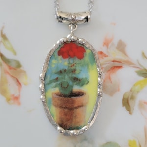 May include: Silver-tone necklace with an oval pendant featuring a painting of a red flower in a brown pot. The background of the painting is yellow and blue. The pendant is framed with a silver beaded edge.