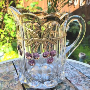 May include: A clear glass pitcher with a gold rim and handle, adorned with gold leaf and cherry accents. The pitcher features a decorative design and is suitable for serving beverages. The pitcher is approximately 8 inches tall.