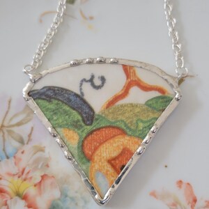 May include: A fan-shaped pendant necklace featuring a colorful floral design on a white background. The pendant is framed in silver and hangs from a delicate silver chain. The design includes orange, green, and blue hues.