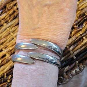 May include: A silver-toned cuff bracelet with a curved design. The bracelet wraps around the wrist, featuring smooth, polished metal. The bracelet is shown on a person's wrist, set against a textured, woven background.