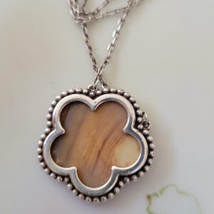 May include: A silver necklace with a clover-shaped pendant. The pendant has a light brown, patterned center and a beaded silver border. The chain is also silver and has a delicate design. The necklace is displayed on a white surface.